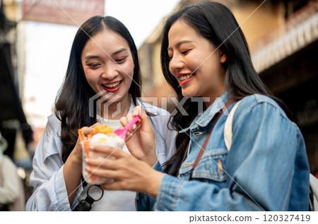 Two pretty, joyful Asian female tourists enjoy street food at a market in Thailand, eating ice cream Two pretty, joyful Asian female tourists enjoy street food at a market in Thailand, eating ice cream 120327419