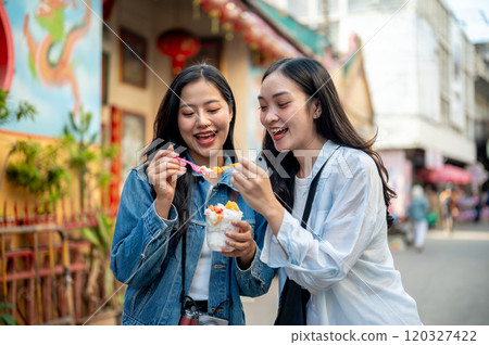 Two pretty, joyful Asian female tourists enjoy street food at a market in Thailand, eating ice cream Two pretty, joyful Asian female tourists enjoy street food at a market in Thailand, eating ice cream 120327422