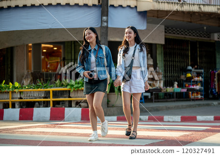 Two joyful young Asian women cross a crosswalk together while strolling through a city in Thailand. 120327491