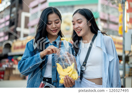 Two happy Asian female tourists enjoy eating while exploring a street food market. Two happy Asian female tourists enjoy eating while exploring a street food market. 120327495