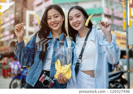Two happy Asian female tourists enjoy eating while exploring a street food market. 120327496