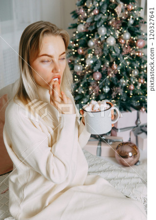 Blonde in a bright Christmas interior with a cup of hot drink with marshmallows. New Year's interior in pink tones. The winter holidays period. Blonde in a bright Christmas interior with a cup of hot drink with marshmallows. New Year's interior in pink tones. The winter holidays period. 120327641