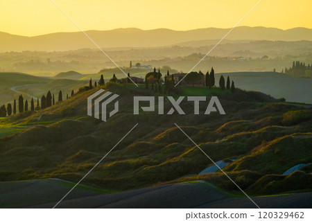 Sunset over rolling Tuscan hills with cypress trees and distant village 120329462