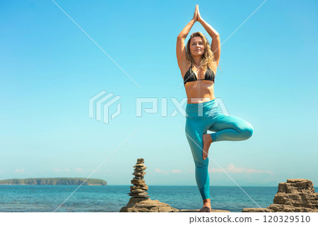 Yoga woman standing in vrikshasana tree pose hand namaste on cliff at sea sky horizon 120329510