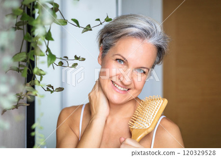 Beautiful middle-aged woman smiling and combing her gray hair in front of the mirror in the bathroom 120329528