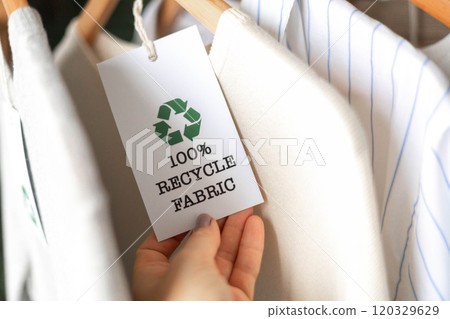 Female hands hold a label from white knitted clothing, on which it says 100 percent recycle fabric Female hands hold a label from white knitted clothing, on which it says 100 percent recycle fabric 120329629
