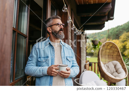 Man is enjoying cup of warm tea, coffee, having relaxing moment at home, standing on patio and looking at autumn nature around him. 120330467