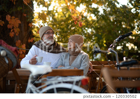 Elderly married couple sitting on a coffee shop terrace on beautiful autumn day, enjoying cup of coffee in the autumn atmosphere. 120330502