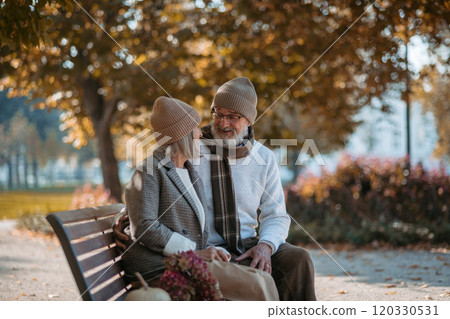 Portrait of beautiful senior couple sitting on bench in autumn park. Elderly husband and wife hugging, enjoying fall atmosphere. 120330531