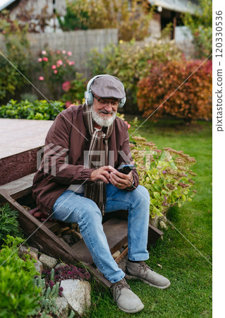 Older man with a gray beard having moment for himself, listening music. Senior wearing headphones and playing music from phone. Older man with a gray beard having moment for himself, listening music. Senior wearing headphones and playing music from phone. 120330536