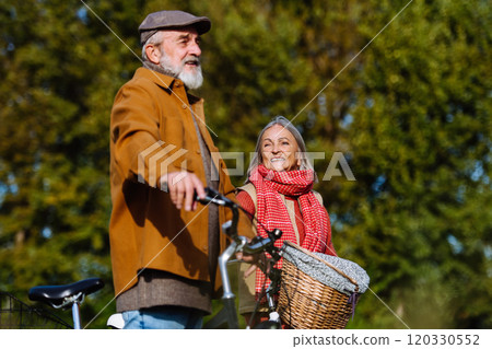Lovely elderly couple on a walk in the forest, pushing their bikes side by side. Seniors in love on stroll through autumn nature, enjoying a peaceful moment together. 120330552