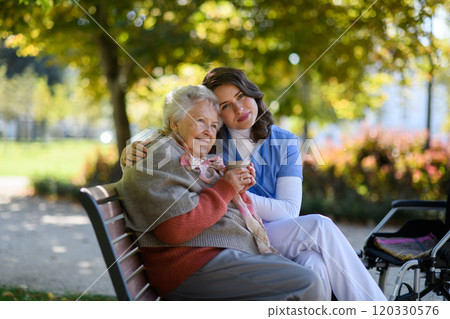 Elderly woman enjoying cup of coffee with her nurse in the park during warm autumn day. Young caregiver spending time with senior woman. 120330576