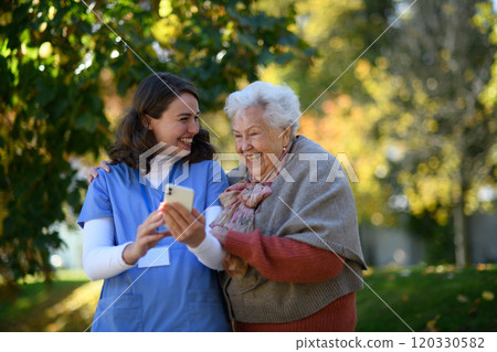 Portrait of nurse and elderly woman on walk in the park during warm autumn day. Young caregiver looking through photos of senior patient's family on a phone. 120330582