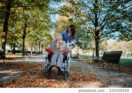 Disabled elderly woman in wheelchair on walk in the park during warm autumn day. Young nurse pushing wheelchair, talking with senior woman. Disabled elderly woman in wheelchair on walk in the park during warm autumn day. Young nurse pushing wheelchair, talking with senior woman. 120330589