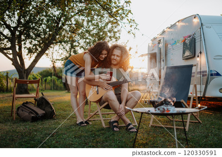 Couple sitting by campfire in the evening, enjoying peaceful moment. Man playing guitar and singing song to his girlfriend. Camping trip for young people. Couple sitting by campfire in the evening, enjoying peaceful moment. Man playing guitar and singing song to his girlfriend. Camping trip for young people. 120330591