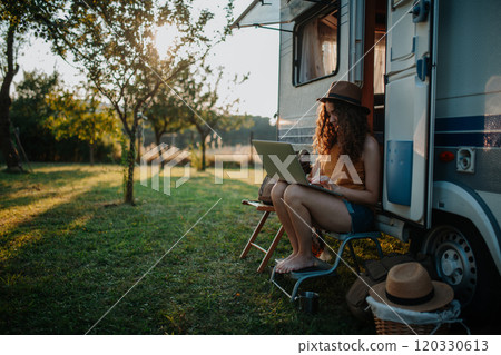 Young beautiful woman is on a camping trip in nature, sitting in front of caravan with notebook on knees. Caravan as homeoffice, working remotely. 120330613