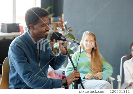 Side view portrait of smiling African American man speaking to microphone while recording podcast with students in art club, copy space 120331511