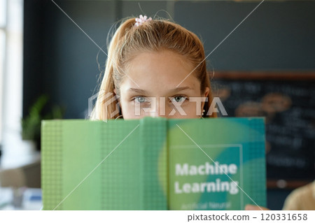 Front view portrait of cute schoolgirl peeking out holding open book on machine learning in IT class at school Front view portrait of cute schoolgirl peeking out holding open book on machine learning in IT class at school 120331658