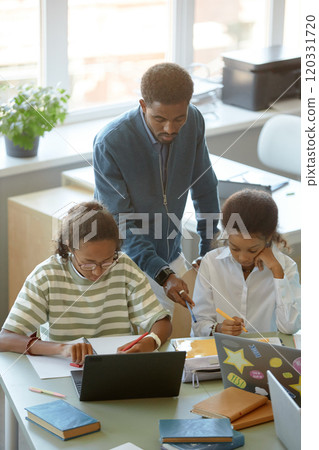 Vertical high angle shot of African American male teacher helping two students in sunlit classroom Vertical high angle shot of African American male teacher helping two students in sunlit classroom 120331720