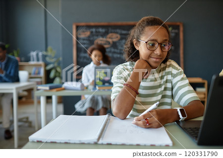 Front view portrait of smiling African American teenage boy wearing glasses and using laptop while studying in school classroom copy space 120331790