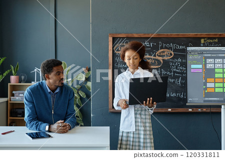 Minimal front view of young African American girl holding laptop while giving presentation in school classroom with teacher watching copy space 120331811