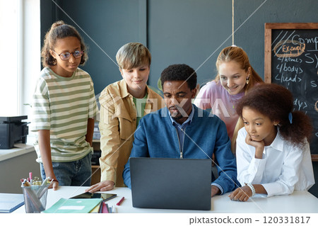 Front view portrait of young male teacher with diverse group of children using computer together in school classroom and smiling Front view portrait of young male teacher with diverse group of children using computer together in school classroom and smiling 120331817