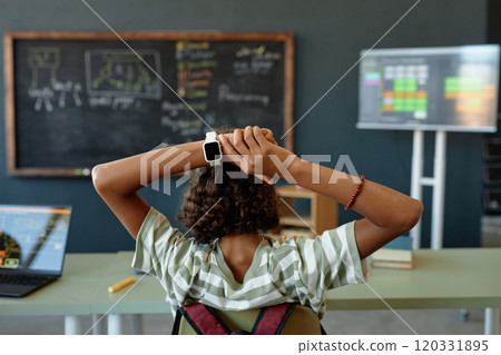 Back view of African American teenage girl stretching while sitting at desk in school classroom with smartwatch in focus wearable tech copy space 120331895