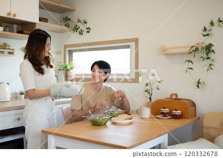 Young woman wearing an oven mitt serving a freshly prepared dish to her husband in a minimalist kitchen Young woman wearing an oven mitt serving a freshly prepared dish to her husband in a minimalist kitchen 120332678
