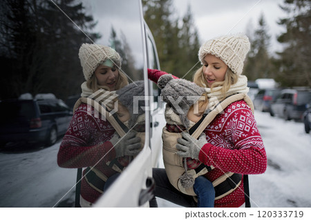 Mother standing in the middle of snowy parking lot with sleeping daughter in baby carrier. Mom and girl enjoying winter holiday in the mountains. Mother standing in the middle of snowy parking lot with sleeping daughter in baby carrier. Mom and girl enjoying winter holiday in the mountains. 120333719