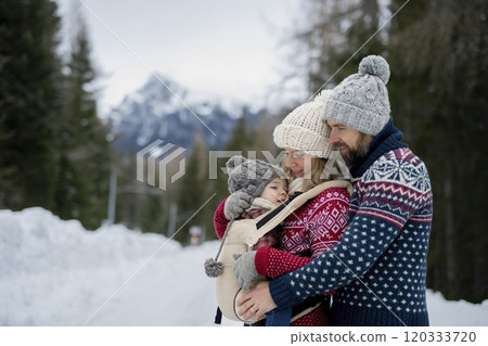 Young family is enjoying winter holiday in the mountains, mother carry small girl in carrier while walking through the snowy forest. 120333720
