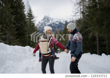 Young family is enjoying winter holiday in the mountains, mother carry small girl in carrier while walking through the snowy forest. 120333721