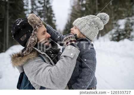 Father standing in the middle of snowy forest holding young son in arms. Dad and boy enjoying winter holiday in the mountains. 120333727