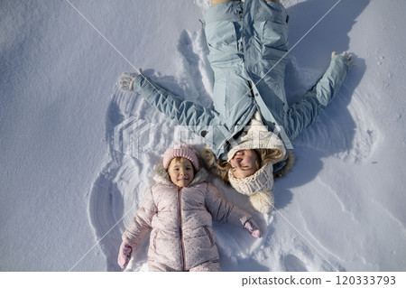 Portrait of small girl and mom making snow angels, enjoying winter holiday in the mountains. Girl lying and playing in snow. 120333793