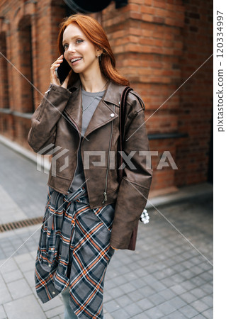 Vertical shot of cute redhead woman happily chatting on phone standing in urban street, exuding confidence and style in trendy attire. Pretty ginger woman chatting on smartphone outdoors. Vertical shot of cute redhead woman happily chatting on phone standing in urban street, exuding confidence and style in trendy attire. Pretty ginger woman chatting on smartphone outdoors. 120334997