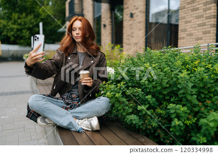 Fashionable young woman with red hair sitting on bench, taking selfie with smartphone while holding cup of coffee. Beautiful redhead female having online video call on mobile phone outdoors. 120334998