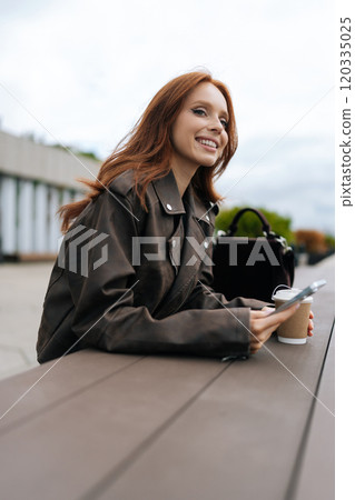 Vertical portrait of cheerfully woman with flowing red hair drinking coffee and checking phone while standing outside by river. Happy redhead female smiling in calm outdoor setting 120335025