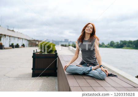 Peaceful redhead woman finding inner peace and balance practicing yoga on tranquil river dock, immersed in nature, inhaling fresh air and embracing sense of calm energy outdoors on cloudy day. Peaceful redhead woman finding inner peace and balance practicing yoga on tranquil river dock, immersed in nature, inhaling fresh air and embracing sense of calm energy outdoors on cloudy day. 120335028