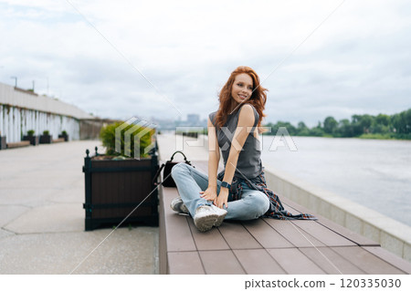 Portrait of happy female finding serenity on river pier, savoring tranquil atmosphere and cloudy sky. Pretty smiling young woman appears at ease, taking respite from urban living outdoors. 120335030