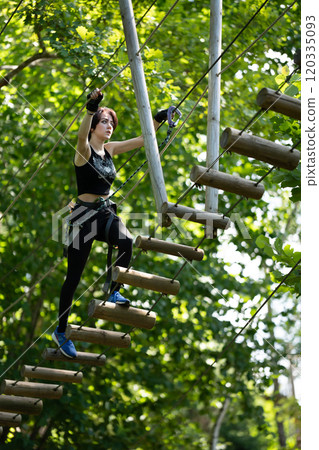 A woman confidently navigates her way across a high ropes course, surrounded by lush, vibrant greenery all around her A woman confidently navigates her way across a high ropes course, surrounded by lush, vibrant greenery all around her 120335093