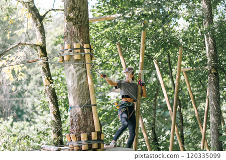An adventurous climber skillfully navigating challenging ropes and wooden structures in a vibrant outdoor setting An adventurous climber skillfully navigating challenging ropes and wooden structures in a vibrant outdoor setting 120335099