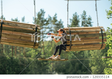 A young woman expertly navigates through a thrilling high ropes course, surrounded by lush trees and warm sunlight 120335114