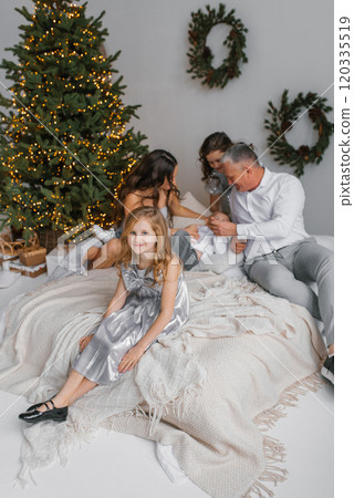 Happy family of five is partying of Christmas tree. Little girl is wearing silver dress and is sitting on bed Happy family of five is partying of Christmas tree. Little girl is wearing silver dress and is sitting on bed 120335519