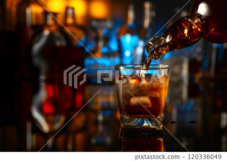 Whiskey is poured from a bottle into a glass with ice. 120336049