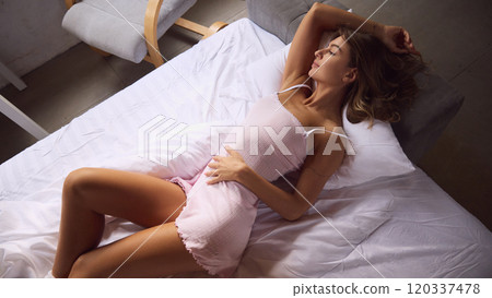 Beautiful woman in soft pink nightgown stretches out on her bed, enjoying relaxed morning in softly lit room. 120337478