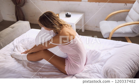 Aerial view of woman in pink nightgown sits on bed, embracing white pillow. Bright room, decorated with indoor plants and soft furnishings. Aerial view of woman in pink nightgown sits on bed, embracing white pillow. Bright room, decorated with indoor plants and soft furnishings. 120337482
