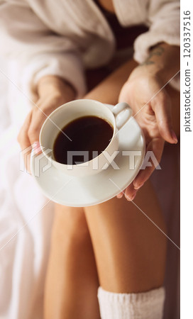 Woman in white robe holding cup of coffee while sitting comfortably on bed with soft blanket. Natural light illuminates cozy bedroom. 120337516