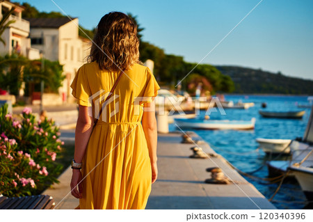 Woman is walking alone on sunny promenade with palm trees and flowers. Female tourist strolls along marina at sunset in coastal town 120340396