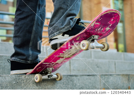 Skateboarder balancing on pink skateboard. Close-up of teenager feet wearing jeans and black sneakers skateboarding at street stairs. Leisure activity and extreme sport concept 120340443