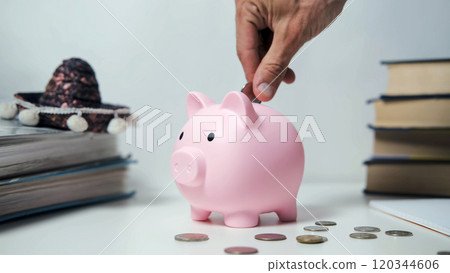 A hand places a coin into a pink piggy bank on a white table surrounded by scattered coins and a crumpled dollar bill, illustrating savings, financial security, and budgeting 120344606