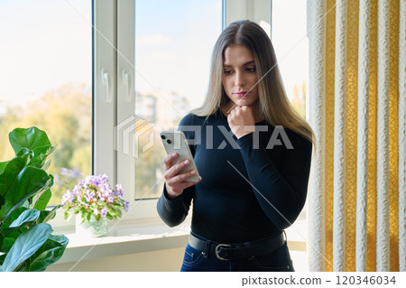 Young serious woman using smartphone standing at home near window 120346034